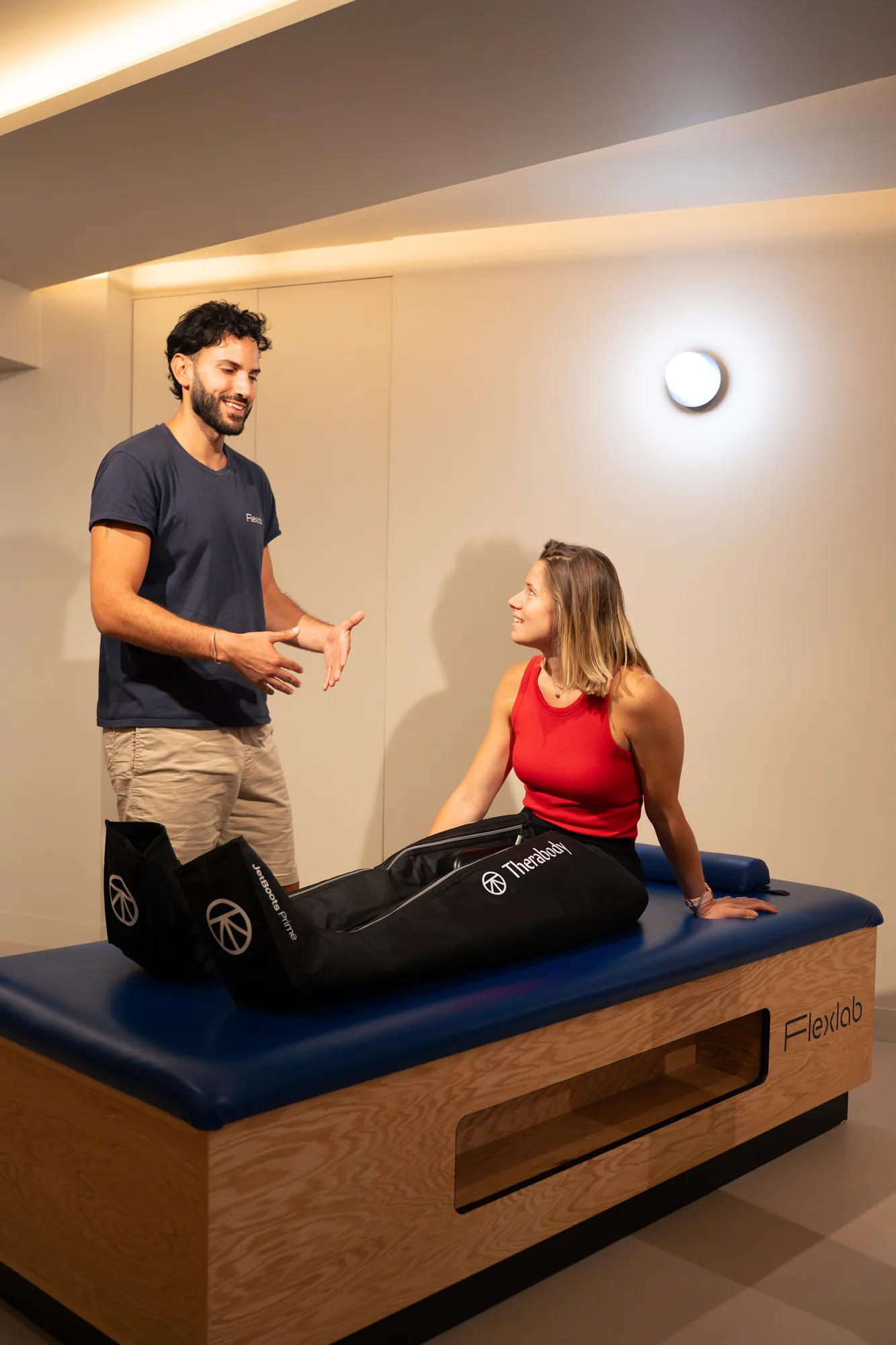 Un flexologue parle avec une cliente assise sur la table, les jambes dans les bottes de pressothérapie